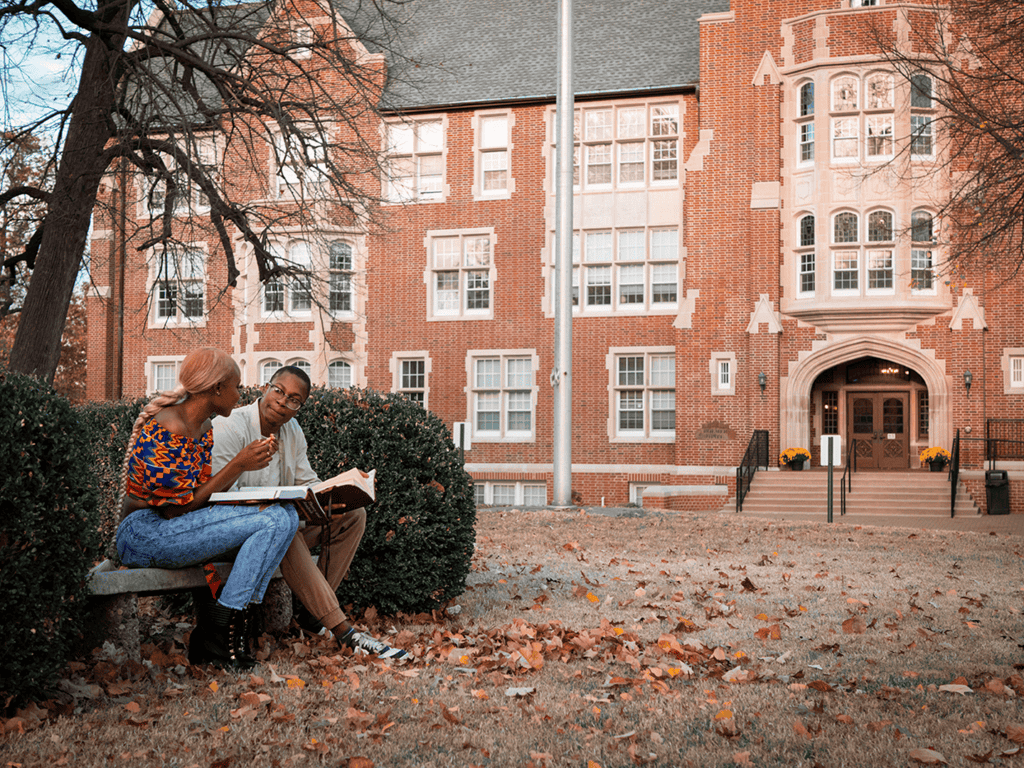Two students studying together on a bench in front of a historic campus building.