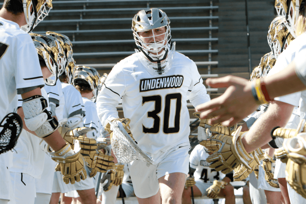 A lacrosse player in Lindenwood University uniform running onto the field with teammates.